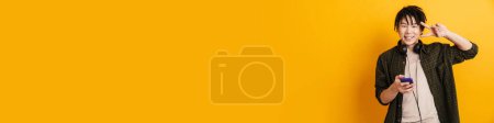 Photo for Smiling young asian man holding mobile phone while standing over yellow background, showing peace gesture - Royalty Free Image