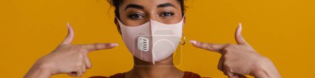 Foto de Black woman looking at camera while pointing finger at her face mask isolated over yellow background - Imagen libre de derechos
