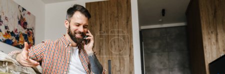 Photo for Smiling brunette bearded man talking on mobile phone while sitting on a floor with laptop computer - Royalty Free Image