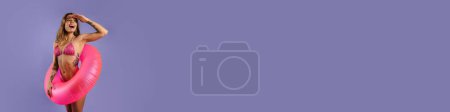Téléchargez les photos : Jeune femme blanche en maillot de bain souriant et tenant bague en caoutchouc isolé sur fond violet - en image libre de droit