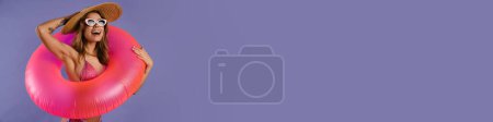Téléchargez les photos : Jeune femme blanche en maillot de bain et lunettes de soleil tenant bague en caoutchouc isolé sur fond violet - en image libre de droit