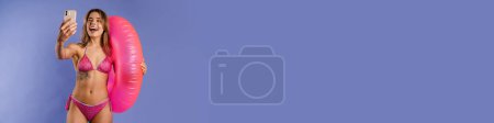 Téléchargez les photos : Jeune femme blanche en maillot de bain tenant anneau en caoutchouc et téléphone cellulaire isolé sur fond violet - en image libre de droit