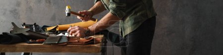 Téléchargez les photos : Artisan barbu concentré travaillant avec le cuir tout en se tenant à table dans l'atelier - en image libre de droit