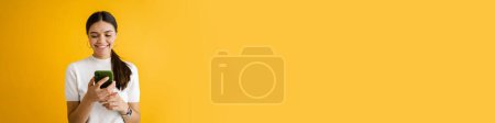 Téléchargez les photos : Jeune femme brune souriante et utilisant un téléphone portable isolé sur fond jaune - en image libre de droit