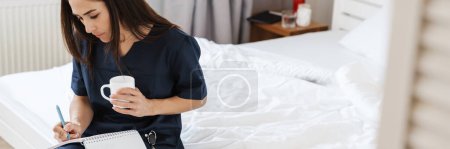 Téléchargez les photos : Une femme brune en uniforme médical bleu assise sur le lit et écrivant quelque chose dans un carnet avec une tasse dans les mains dans la chambre - en image libre de droit