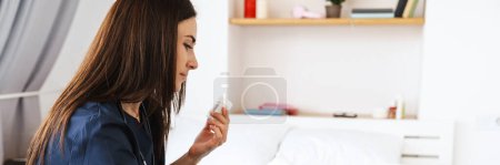 Photo for A side view of brunette woman in blue medical uniform sitting in front of laptop and showing jar of pills to laptop camera in bright room - Royalty Free Image