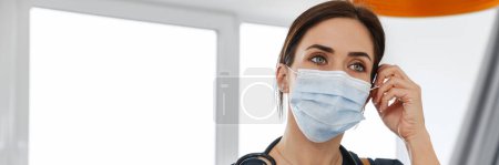 Photo for The woman doctor holding a tablet with papers and a phone in her hands, straightens a mask on her face while standing in a white room - Royalty Free Image