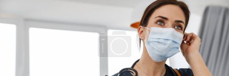 Photo for A woman in a medical uniform with a stethoscope straightening a mask on her face while holding a phone and a tablet with papers in the room - Royalty Free Image