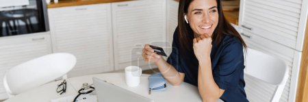 Photo for Smiling woman doctor using credit card and laptop while sitting at home - Royalty Free Image