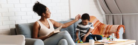 Photo for Black woman reading book while her son playing with toys at home - Royalty Free Image