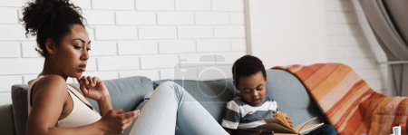 Foto de Mujer negra usando celular mientras su hijo lee libro en casa - Imagen libre de derechos