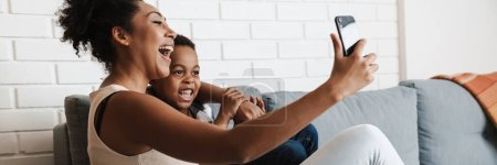 Téléchargez les photos : Femme souriante noire prenant des photos selfie sur son téléphone portable tout en embrassant son fils à la maison - en image libre de droit