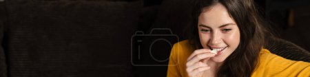 Téléchargez les photos : Sourire jeune femme brune manger du pop-corn tout en étant assis sur un canapé à la maison - en image libre de droit