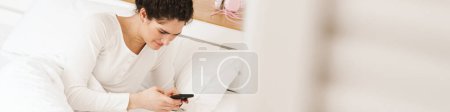 Téléchargez les photos : La femme allongée sur le lit blanc en pyjama et regardant le téléphone dans la pièce lumineuse - en image libre de droit