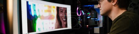 Téléchargez les photos : Vue arrière d'un jeune homme blanc monteur vidéo assis devant un grand écran travaillant sur un montage vidéo à l'intérieur - en image libre de droit