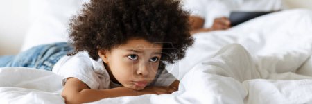 Foto de Niño negro frunciendo el ceño mientras su padre usa la tableta en la cama en casa - Imagen libre de derechos
