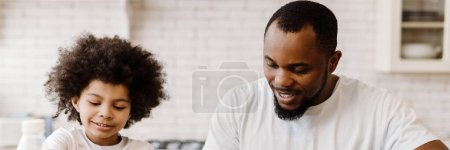 Téléchargez les photos : Père et fils noirs souriant tout en prenant le petit déjeuner à la maison - en image libre de droit