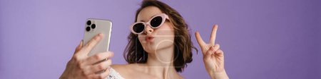 Photo for Happy young white woman holding mobile phone standing over violet background, taking a selfie, wearing sunglasses - Royalty Free Image