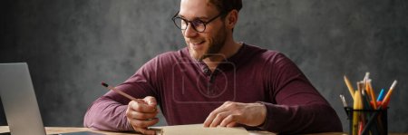 Téléchargez les photos : Un homme souriant assis à une table devant des papiers tout en regardant l'écran de l'ordinateur portable dans le studio - en image libre de droit