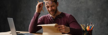 Photo for The smiling man reading papers with raising glasses while sitting at table in the studio - Royalty Free Image