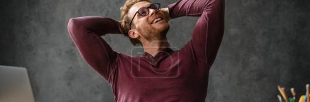 Photo for The smiling man in glasses looking to the side with raising hands while sitting at a table in the studio - Royalty Free Image