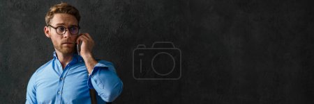 Photo for The upset man in a blue shirt and glasses with a bag on his shoulder talking on the phone while wrinkling his forehead in the studio - Royalty Free Image