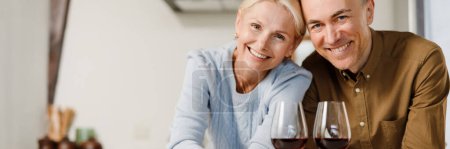Photo for Smiling middle aged couple standing together at the kitchen drinking red wine at home - Royalty Free Image