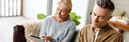 Photo for Smiling middle aged white couple relaxing on a couch at home, relaxing, using tablet, writing in notebook - Royalty Free Image