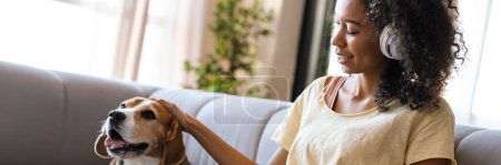 Téléchargez les photos : Jeune femme africaine en tenue décontractée avec chiot beagle à la maison, assise sur un canapé, utilisant une tablette dans un casque - en image libre de droit