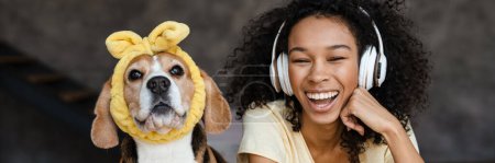 Téléchargez les photos : Jeune femme africaine heureuse en tenue décontractée avec beagle chiot à la maison, se détendre sur le lit dans des écouteurs riant - en image libre de droit