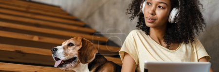 Foto de Joven mujer africana sonriente en ropa casual con perrito beagle en casa, sentado en las escaleras, sosteniendo la tableta, usando auriculares - Imagen libre de derechos