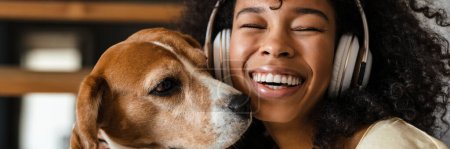 Téléchargez les photos : Jeune femme africaine en tenue décontractée avec beagle chiot à la maison, assis sur les escaliers dans les écouteurs, cose up, jouer avec le chien - en image libre de droit