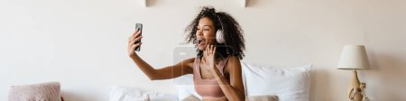 Photo for Smiling mid aged african woman listening to music with headphones while relaxing on bed at home, taking a selfie or on a video call - Royalty Free Image