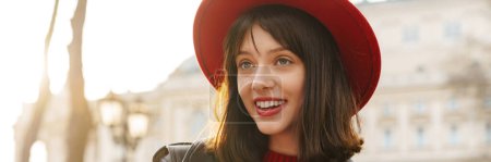 Photo for Close up of a smiling young stylish brunette white woman student wearing hat and leather jacket with short hair standing on a street carrying copybooks - Royalty Free Image