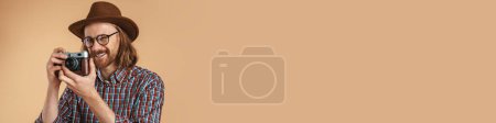 Téléchargez les photos : Rire gingembre homme portant des lunettes et un chapeau à l'aide d'une caméra rétro isolé sur fond beige - en image libre de droit