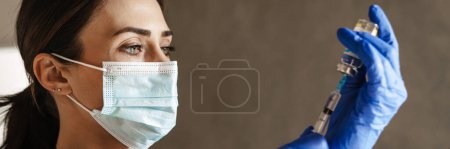 Photo for Confident mid aged brunette woman doctor wearing mask and gloves holding syringe indoors, close up - Royalty Free Image