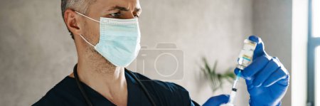 Photo for Mid aged male doctor wearing protective mask holding a syringe with vaccine - Royalty Free Image