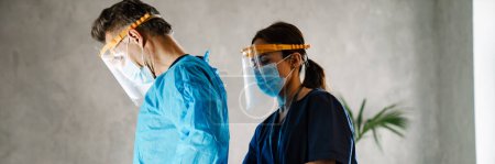 Photo for Two medical workers dressing up in uniform standing indoors, wearing robes and masks - Royalty Free Image