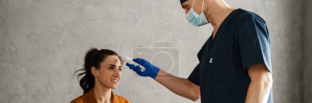 Photo for Woman patient and man doctor sitting in the cabinet during medical examination, doctor measuring temperature - Royalty Free Image