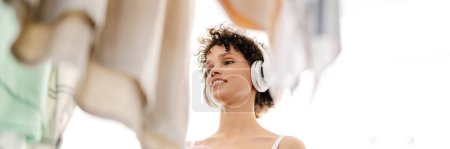 Photo for Young white smiling woman chooses clothes in the wardrobe closet at home, wearing wireless headphones, holding mobile phone - Royalty Free Image