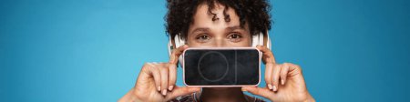 Téléchargez les photos : Jeune femme brune bouclée portant des écouteurs montrant le téléphone cellulaire isolé fond bleu blanc - en image libre de droit