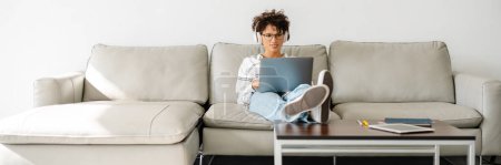 Foto de Mujer rizada joven en los auriculares usando el ordenador portátil mientras está sentado en el sofá en casa - Imagen libre de derechos