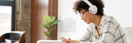 Téléchargez les photos : Jeune femme bouclée dans un casque à l'aide d'un ordinateur portable et d'un téléphone portable assis sur le canapé à la maison - en image libre de droit