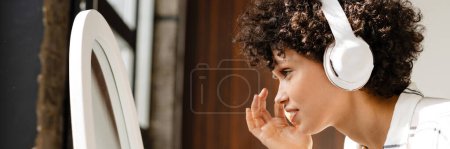 Téléchargez les photos : Jeune femme bouclée écoutant de la musique avec des écouteurs tout en regardant miroir à la maison - en image libre de droit
