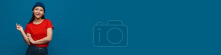 Téléchargez les photos : Asiatique adolescent fille en chapeau souriant et regardant caméra isolé sur fond bleu - en image libre de droit