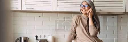 Photo for Mature smiling woman talking on cellphone while standing in kitchen at home - Royalty Free Image