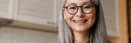Téléchargez les photos : Mature femme grise souriant tout en travaillant avec un ordinateur portable dans la cuisine à la maison - en image libre de droit