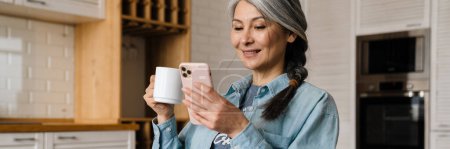 Photo for Mature grey woman smiling and using cellphone while drinking coffee at home - Royalty Free Image
