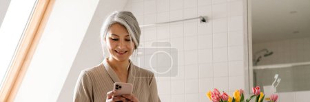 Foto de Mujer gris madura sonriendo mientras usa el teléfono móvil en el baño en casa - Imagen libre de derechos