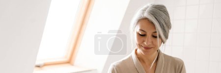 Foto de Mujer gris madura sonriendo mientras usa el teléfono móvil en el baño en casa - Imagen libre de derechos